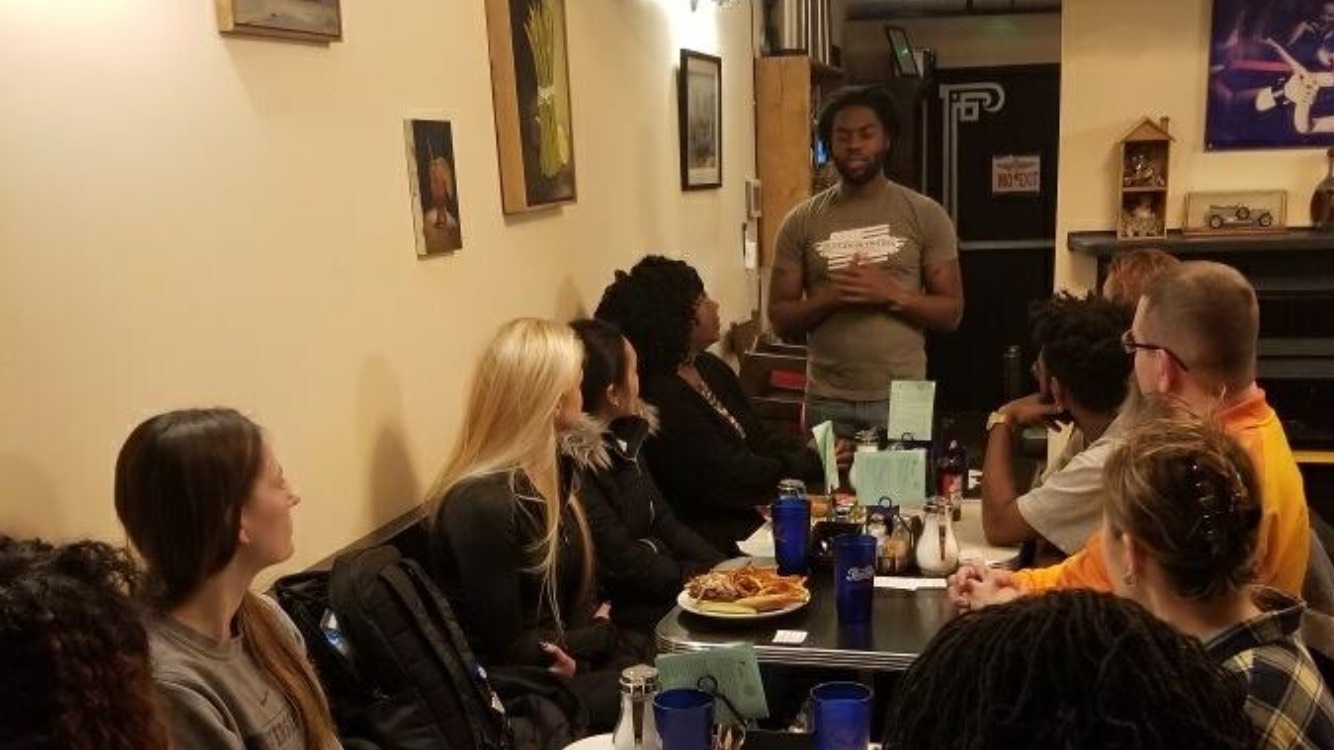 Mack McLelllan, an African American Man, speaks to a group of people sitting at dining tables.
