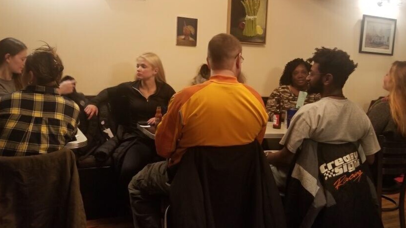  group of people sitting at dining tables in a restaurant. Participants are a mixture of ages -- ranging from early 20s to late 40s, present as different races and genders. They are engaged in small group conversations.